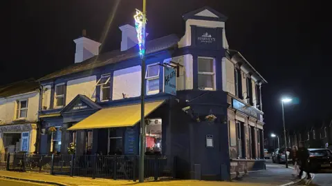 A blue and white pub on a corner. There are signs which say "The Heath Tavern".