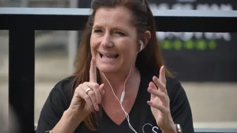 Carol Kyle, BSL interpreter, signs with both her fingers up, she wears white headphones and is mouthing something.