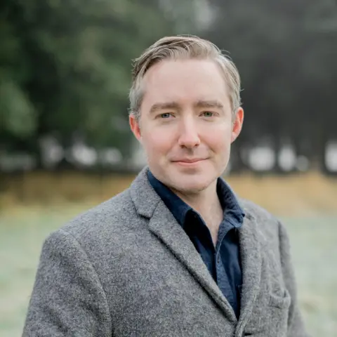 Lonán Ó Briain Lonán Ó Briain standing outside wearing a grey jacket and blue shirt