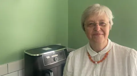 A woman stands to the right of the photo, smiling at the camera. She has short white hair and is wearing glasses. She is wearing a white blouse with an orange beaded necklace. Behind her is a wall painted light green, some white kitchen tiles and an air fryer appliance.