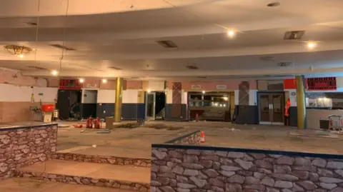 LDRS The space is wide and mostly empty, with a rough, unfinished floor and scattered debris visible across the ground. In the foreground, there is a low, raised platform faced with stone‑patterned panels.
The ceiling is damaged and partially stripped, with missing panels, exposed wiring, and recessed light fixtures hanging down. Several lights are switched on. Structural columns painted in muted colours run through the space, dividing the room into sections.
Along the back wall, there are multiple doorways and openings, some leading into darker adjoining rooms. Tools, equipment, and construction materials are scattered around.