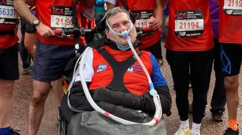 Motor Neurone Disease campaigner Sam Perkins in a wheelchair and wearing ventilation tubes connected to his nose. He is wearing a red T-shirt and surrounding by people also wearing red t-shirts with numbered race bibs.