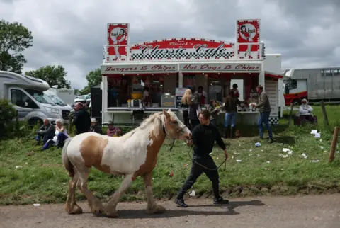 ADAM VAUGHAN/EPA/Shutterstock A white and brown horse is led past a food truck by a man. The food stall is serving burgers and hot dogs.