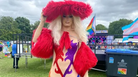 Michelle Alexandria with blonde hair and a large red boa hat. She is wearing a cream, red and purple one