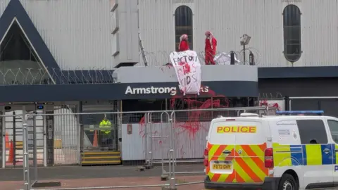 The two protesters on the gatehouse of Armstrong Works. They are holding a sign which reads, this factory kills kids. A police van is parked outside the building.