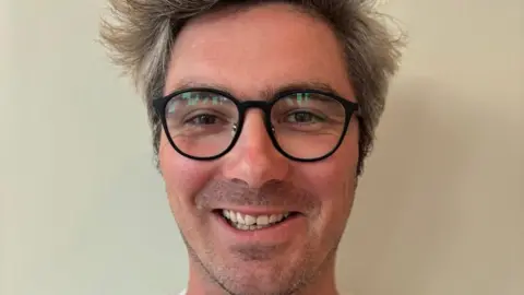 Tom Roberts Tom Roberts is Caucasian, has short greying blonde hair and is wearing dark rimmed glasses. He is smiling at the camera.