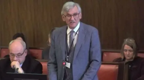 Warwickshire County Council Conservative Councillor Peter Butlin, Warwickshire County Council's Portfolio Holder for Finance, stands to speak in the council chamber, There are red leather and wooden benches behind him and he is wearing a grey suit, tie and waistcoat.