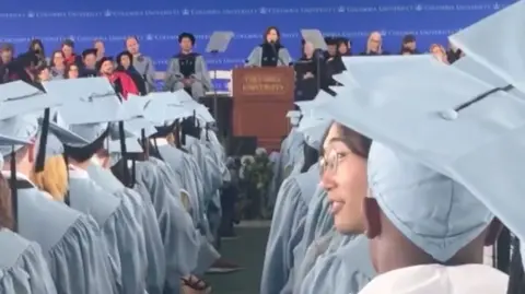 A woman looks over her left shoulder among a crowd of graduates wearing baby blue robes and graduation caps.