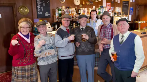 The cast of Still Game all looking at the camera. There are six people standing in a row in front of a bar. A seventh person, wearing a blue shirt, is behind the bar.