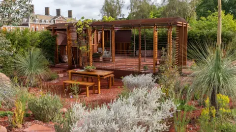 RHS / Sarah Cuttle Wooden garden structure with seating and plants around it, set in front of a large brick building.