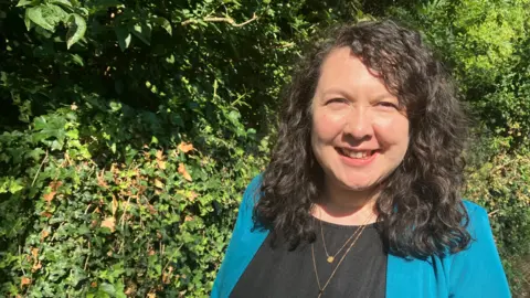 Amy Holmes/BBC Victoria Collins MP with long dark hair standing outdoors in bright sunlight in front of a dense hedge covered in green ivy and leaves. She is wearing a teal jacket over a black top and a necklace, with the foliage filling the background.