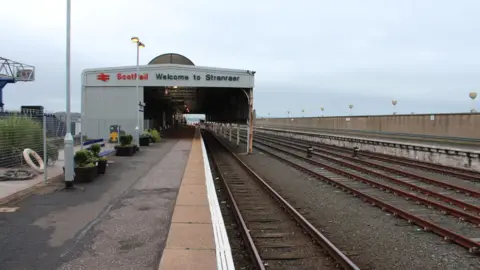 Billy McCrorie Stranraer railway station with tracks running to a grey structure over one set of tracks and a sign saying Welcome to Stranraer