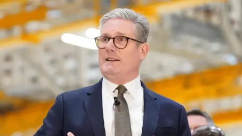 Keir Starmer speking to staff of an aerospace site. He is wearing a blue suit with a grey tie. He is standing in front of an orange and white blurred background