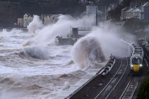 Getty Images A train travelling along the coastal railway at Dawlish beside the newly built sea wall, with a pedestrian walkway and houses on the right and cliffs rising behind them.