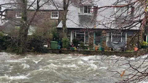 BBC A house built next to the Afon Glaslyn has what appears to be a low wall around it. The river level is close to the top of the wall with areas of white water and waves seen.