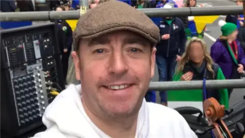 Facebook In a selfie, Ben Gunnery is wearing a brown flat cap and a white hoodie. He appears to be on a stage above a crowd with metal railings. There is a black box behind him with various dials and buttons. At the bottom of the photo you can see that he is holding a brown violin. 