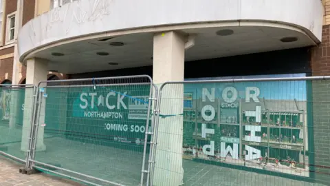 The outside of a former shopping centre, with metal fencing outside and pictures and wording behind it. There are large white columns holding up what appears to be a first-floor balcony.