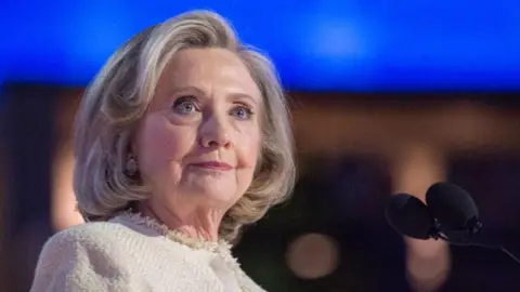 Hillary Clinton has short blonde hair which is curled in at the bottom. She has a cream top on and is wearing makeup as she stands in front of a black microphone.