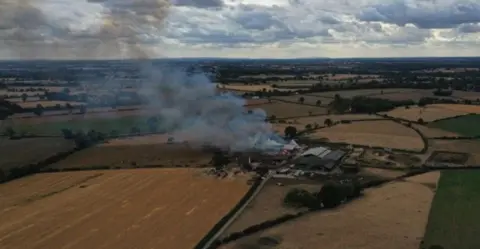 Eliis Dickens An aerial image of an agricultural farm in the middle of many fields is on fire. the flames are large and orange with grey smoke billowing from them and into the air