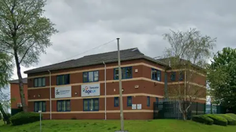 Red brick building with blue window frames surrounded by green grass. There is an Age UK sign on the front of the building.