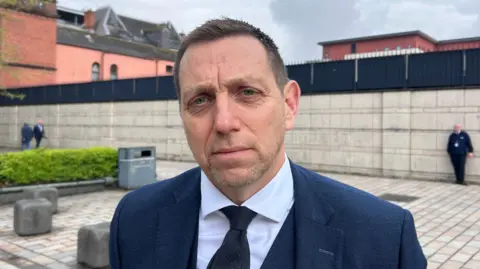 A man with short brown hair, looking at the camera. He is wearing a navy suit, navy tie and white shirt. 