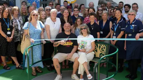 John Fairhall/BBC The parents sitting in front of the ribbon, Nathan is holding the scissors. There are several members of NHS staff standing behind them 