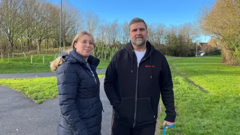 Trish and Richard Mitchell are out walking their dogs (who are not in the image) they are pictured wrapped up in warm coats. Richard can be seen holding a dog lead in his hand. Behind them is a tarmacked footpath, tree saplings and grass area. 