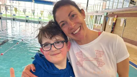 Liz Hardy Ben, standing next to his swimming teacher, Laura Ager, by the side of a swimming pool. He is wearing glasses, has a blue top on, dark short hair and is putting his thumb up, he is smiling, Laura, has on a white top, is looking at the camera and has her head tilted to the right. A pool is behind them.