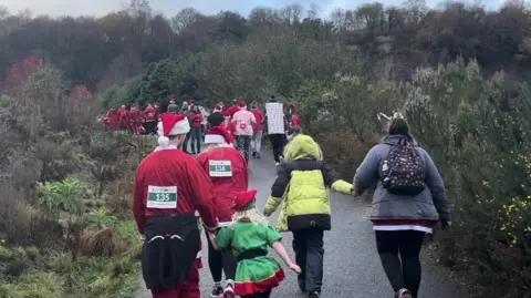 A group of people run up a hill surrounded by large bushes. Many are wearing santa outfits and have running numbers taped to their back. Two people on the right are wearing coats. 