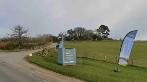 Google A sign saying Tregoad Holiday Park on a patch of grass by the side of a road with two blue flags and a T-junction leading to woodland.