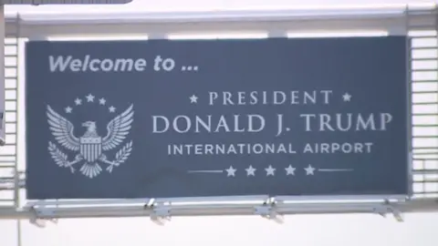 Image shows a sign saying "Welcome to President Donald J Trump International Airport"