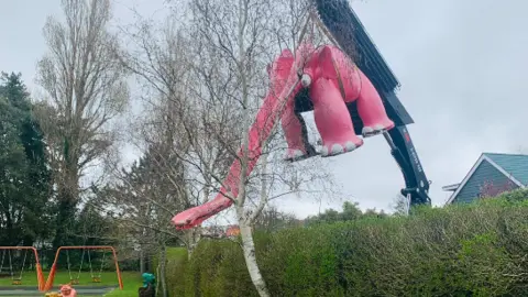 Chris Orkizewski A giant pink, slightly discoloured pink elephant slide being lifted out of a park over a hedge via winch.
