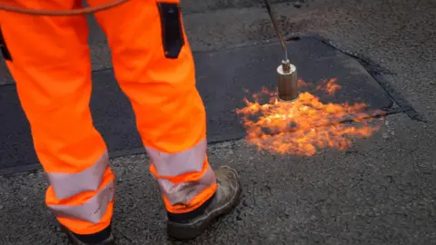 PA Media A workers legs in high vis orange trousers shown as a tarmac patch repair is sealed using a flame