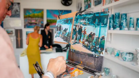Henley Royal Regatta / Row360 A man paints a picture of Henley Regatta in a shop while a woman is talking to another woman behind the counter.