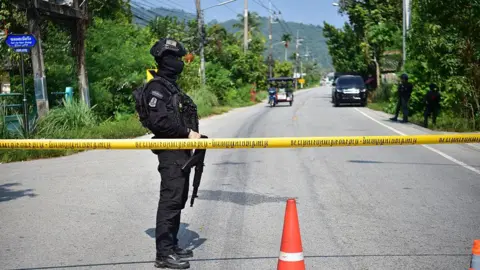 AFP via Getty Images A soldier completely clad in a black uniform and holding a gun stands at a yellow police tape which stretches over a road. A red traffic cone is in the foreground.