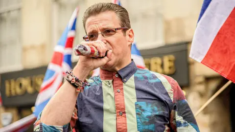 Getty Images Alistair McConnachie, who has short brown hair, speaks into a microphone. He is wearing a union flag coloured shirt. 