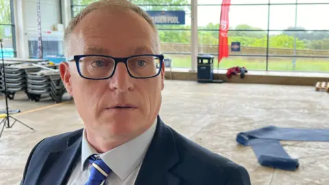 BBC/RICHARD EDWARDS North Yorkshire Council's Conservative deputy leader Gareth Dadd is looking to the right of the camera. He is wearing spectacles, a blue jacket, a pale shirt and a blue-and-white tie.