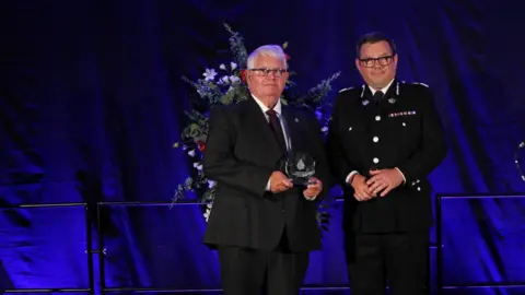 Dyfed-Powys Police Glan Thomas holding his trophy on the stage alongside a sergeant on stage. Both are looking at the camera. Behind them is a large flower arrangement. Glan, on the left, is wearing a black and white suit with a maroon tie. On the right, the sergeant is wearing a black officer's suit with a white shirt and black tie.