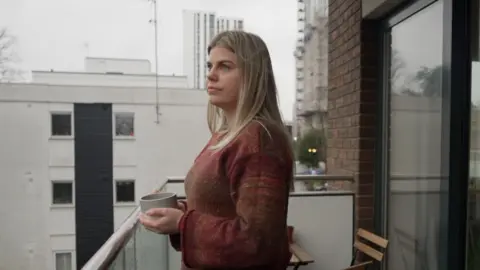 Deryn Harris is standing on her balcony holding a grey mug and looking out over the city