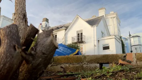 A white hotel in the background is in focus. Part of its roof is burned out and the windows are smashed. A blurry tree stump and a blue bag full of construction materials is in the foreground.