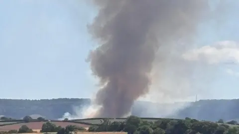 Thick black smoke billows over fields in Devon. It is rising into the air.