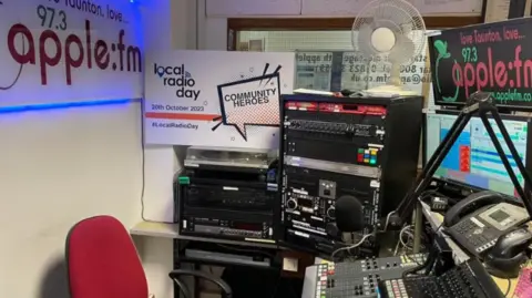 Apple FM A community hospital radio station, featuring a desk full of technical equipment, a computer monitor, and a telephone. There is Apple FM branding all over the space and a fan stacked on top of a large box.