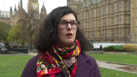 Layla Moran speaks outside Parliament. She wears a purple coat and a red and yellow silk scarf and wears her dark brown, shoulder length hair loose.