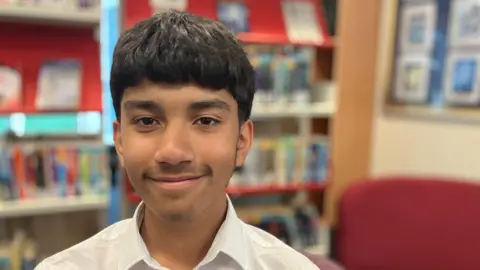 A boy called Navaj is smiling at the camera. He is sat in a library and is wearing a white shirt. He has black hair. 