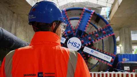 HS2 worker in a tunnel with a tunnel boring machine in front. He is wearhing an orange high-vis jacket and a blue hard hat