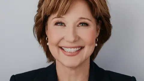 Christy Clark Christy Clark yang tersenyum mengenakan jaket hitam cerdas dan anting-anting putih.