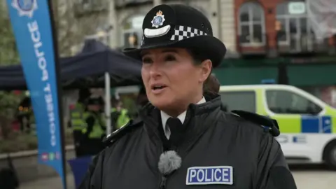 Ch Supt Julie Howe does a TV interview in a street with a police car in the background. She wears police uniform and has brown hair tied back in a bun.