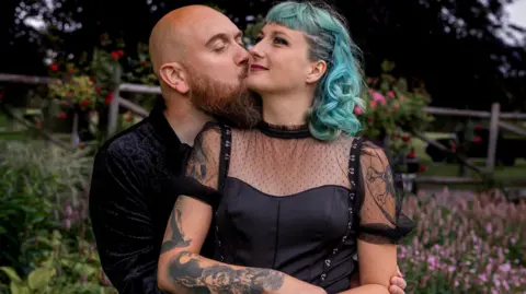 A man with a bald head and long beard has his arms around a woman and is kissing her cheek. The woman has short blue hair and a black dress with sheer material. Behind them are pink and red flowers and a wooden fence