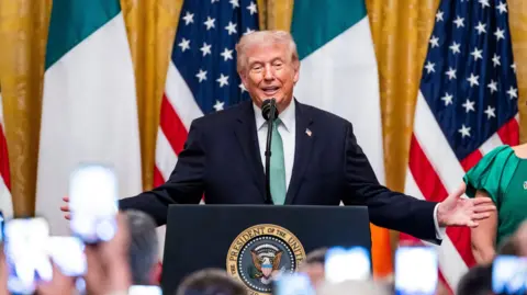 Donald Trump wearing dark suit and green tie. Standing at a podium in front of Irish and American flags. Trump has grey hair.