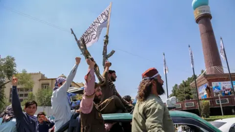 Two men brandishing weapons ride on top of a green car with others around them raising fists